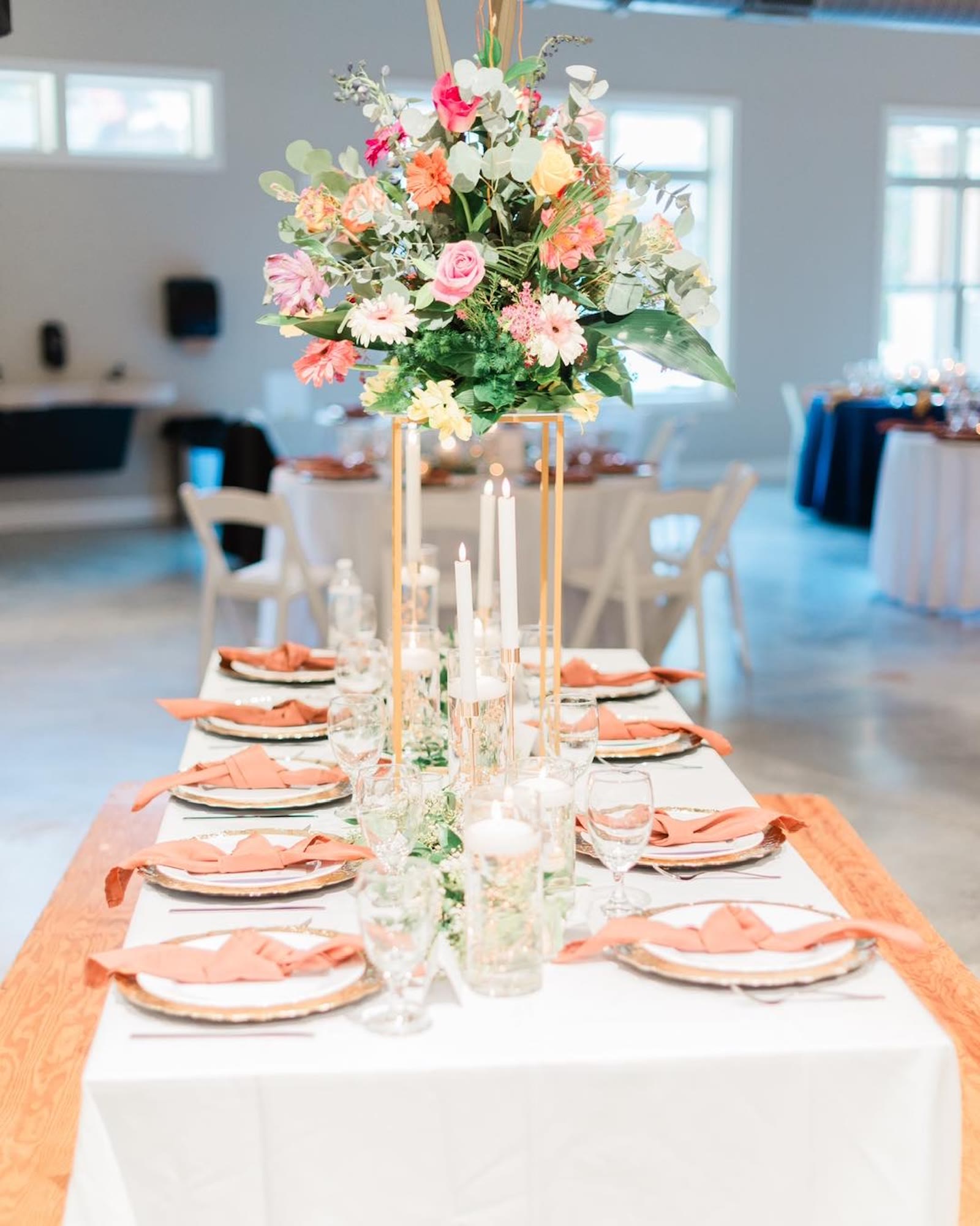 A long banquet table in a bright room, covered with white linen. Terracotta napkins rest on gold-rimmed chargers beside clear glassware. A tall gold stand holds a large arrangement of pink roses, coral and white gerbera daisies, yellow blooms, and eucalyptus. White tapers and floating candles run down the center. Round tables with navy and white cloths sit in the background.
