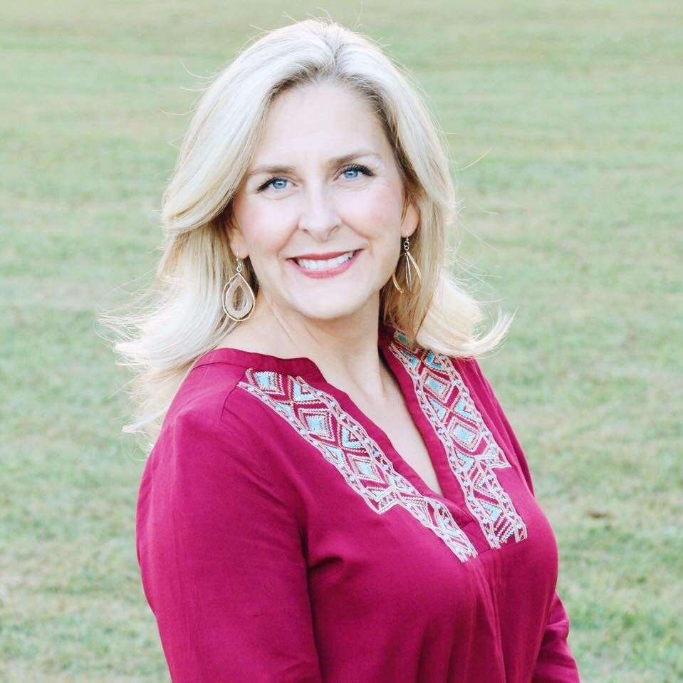 Amy Brown, founder of Graceful Settings, smiling outdoors. She has shoulder-length blonde hair and wears a burgundy top with turquoise and cream embroidery and gold teardrop earrings. The background is a soft green field.