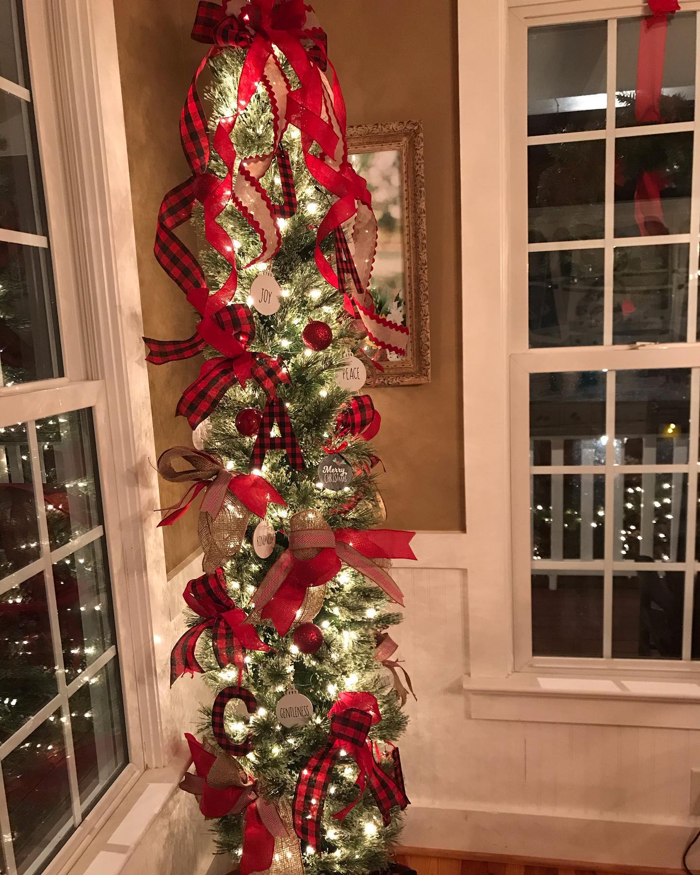 A tall Christmas tree in a corner between white-trimmed windows, covered in white lights. Wide red, black plaid, and burlap ribbons cascade down it. Round ornaments and oval word ornaments are visible, including ones reading Joy, Peace, Merry Christmas, Kindness, and Gentleness, plus a black letter C. A framed picture hangs on the tan wall behind the tree.