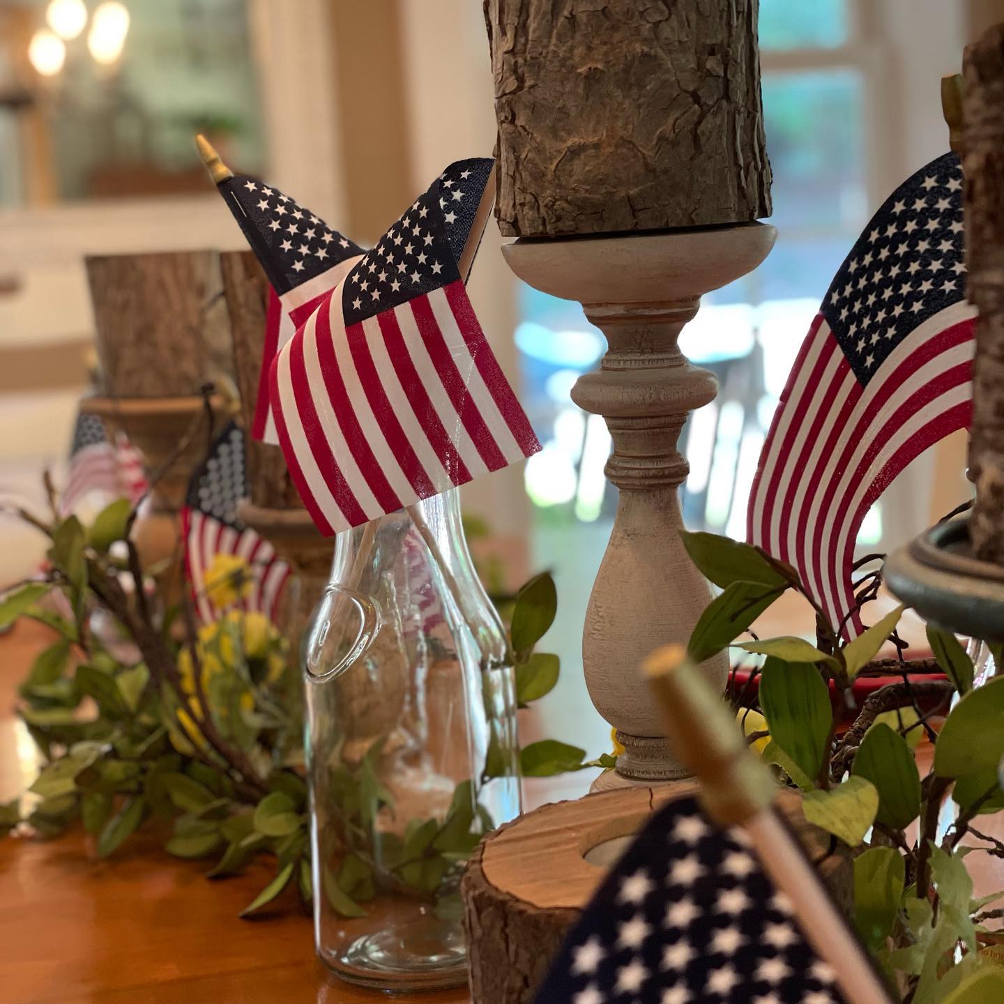 A close view of a wood table set with small American flags on sticks, sprigs of green leaves, a clear glass bottle, and turned wood candlesticks holding pieces of bark. More flags and greenery continue down the table. A bright window is in the background.