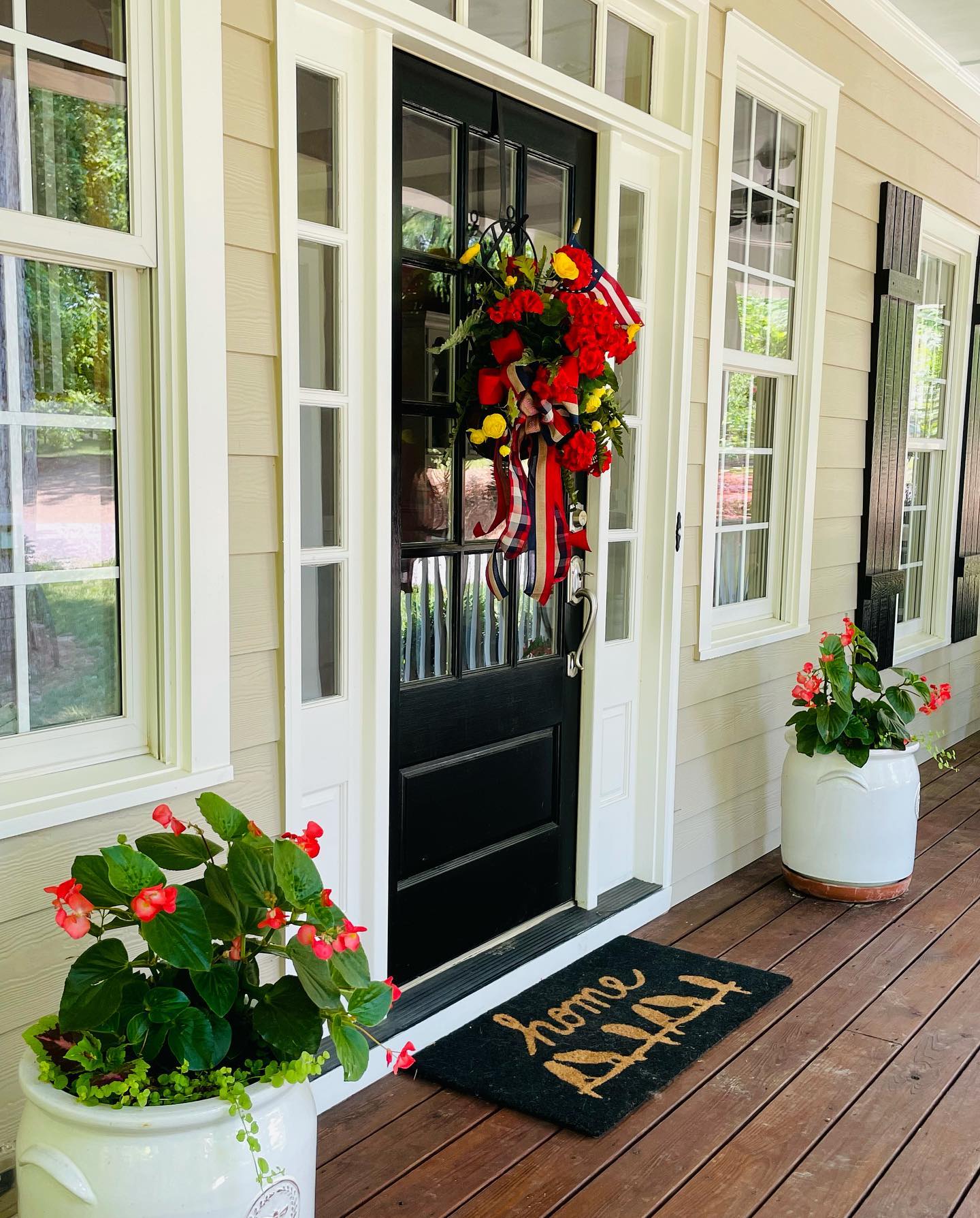 A black front door with glass panes, set in cream siding with white trim. A wreath of red and yellow flowers hangs on the door with long red, white, and navy ribbon. White crocks of red begonias stand on the wood porch on either side. A dark doormat in front of the door reads home.