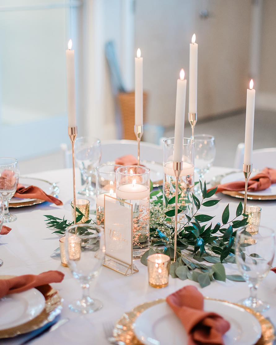 A close view of a tablescape on white linen: tall white tapers in slim gold holders, floating candles in clear glass cylinders, eucalyptus and tiny white blooms, terracotta napkins, gold-rimmed plates, and small gold votive holders. A small gold-framed card stands among the glassware.